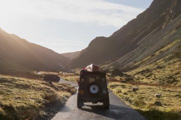 Image of a Land Rover with a canoe on the roof driving up Honister Pass whilst the sun is shining.
