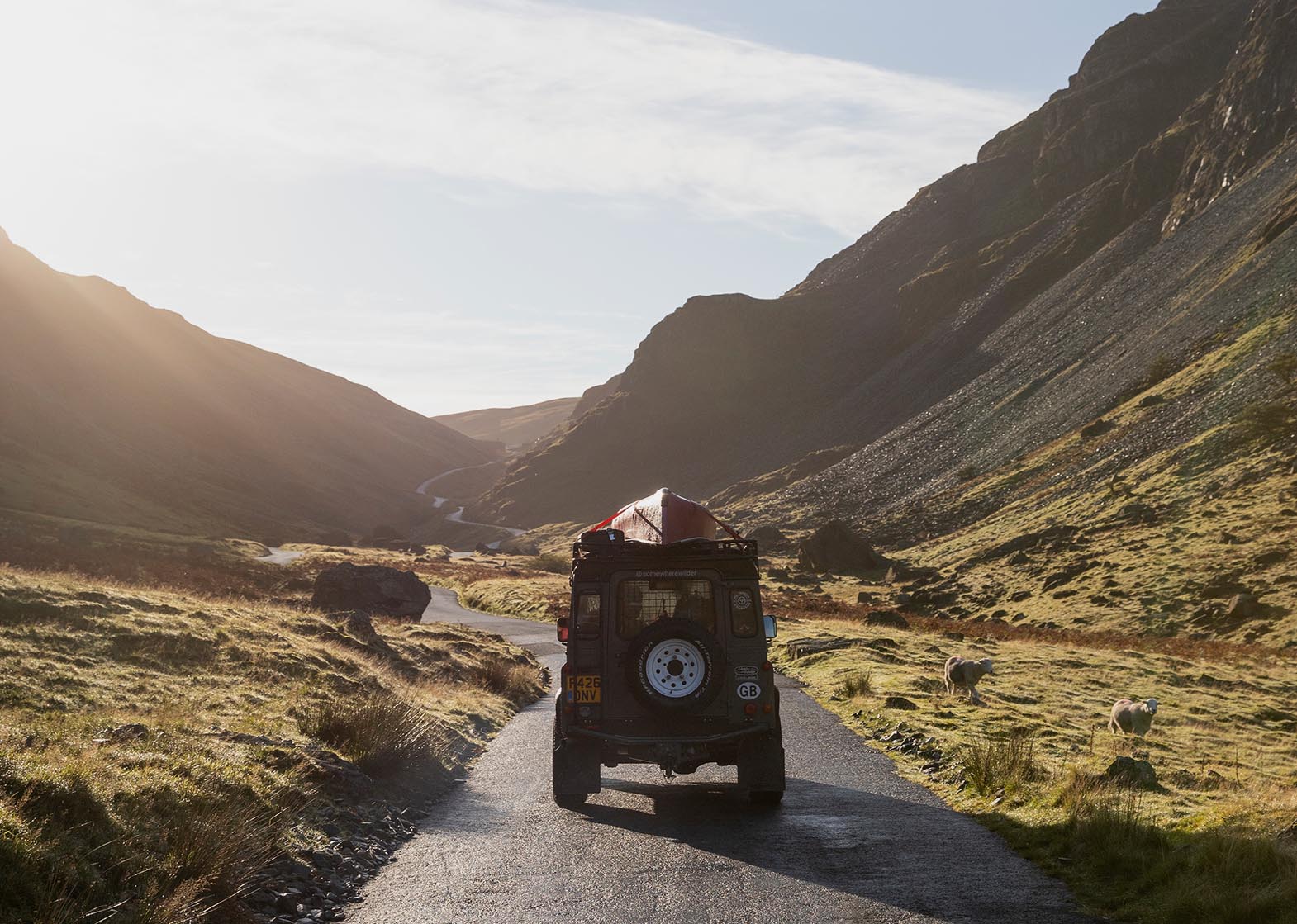 Image of a Land Rover with a canoe on the roof driving up Honister Pass whilst the sun is shining.